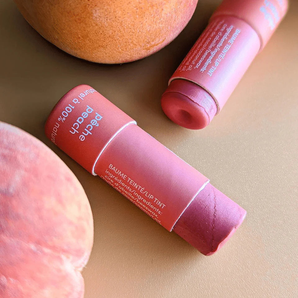 Baume à lèvres teinté Mae Mae à la pêche montré ouvert, posé sur une surface beige avec des pêches fraîches, mettant en valeur la texture crémeuse du baume.

