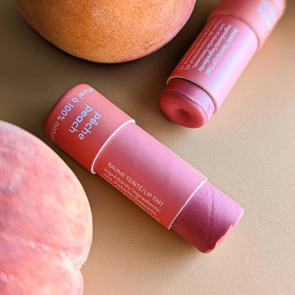 Baume à lèvres teinté Mae Mae à la pêche montré ouvert, posé sur une surface beige avec des pêches fraîches, mettant en valeur la texture crémeuse du baume.
