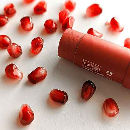 Closed Mae Mae pomegranate lip balm tube lying on its side on a light surface, surrounded by scattered glossy red pomegranate seeds