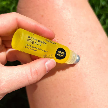 Roll‑on naturel apaise piqûres et chasse‑moustiques Mae Mae tenu à la main en plein air, avec des arbres et un ciel bleu en arrière‑plan, illustrant un usage lors d’activités extérieures.
