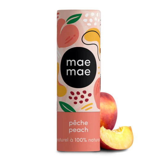 Baume à lèvres teinté Mae Mae à la pêche présenté debout dans un tube illustré aux tons pêche, placé devant une pêche fraîche et un quartier de pêche sur fond blanc.