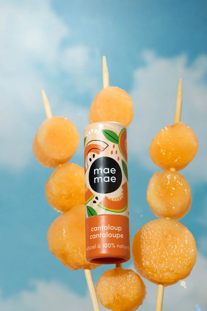Baume à lèvres teinté naturel Mae Mae cantaloup suspendu devant des billes de cantaloup sur fond de ciel bleu
