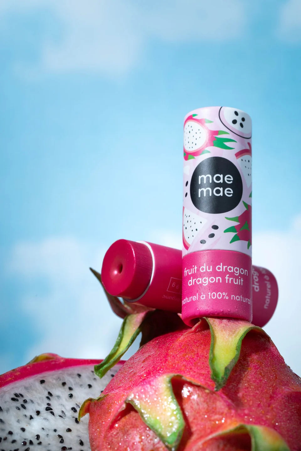 Baume à lèvres teinté Mae Mae au fruit du dragon posé sur des fruits du dragon frais, avec un ciel bleu en arrière‑plan et le tube partiellement ouvert.