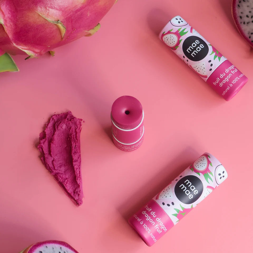 Composition sur fond rose montrant plusieurs baumes à lèvres teintés au fruit du dragon Mae Mae, un bâton ouvert et une trace de texture rose, entourés de morceaux de fruit du dragon.