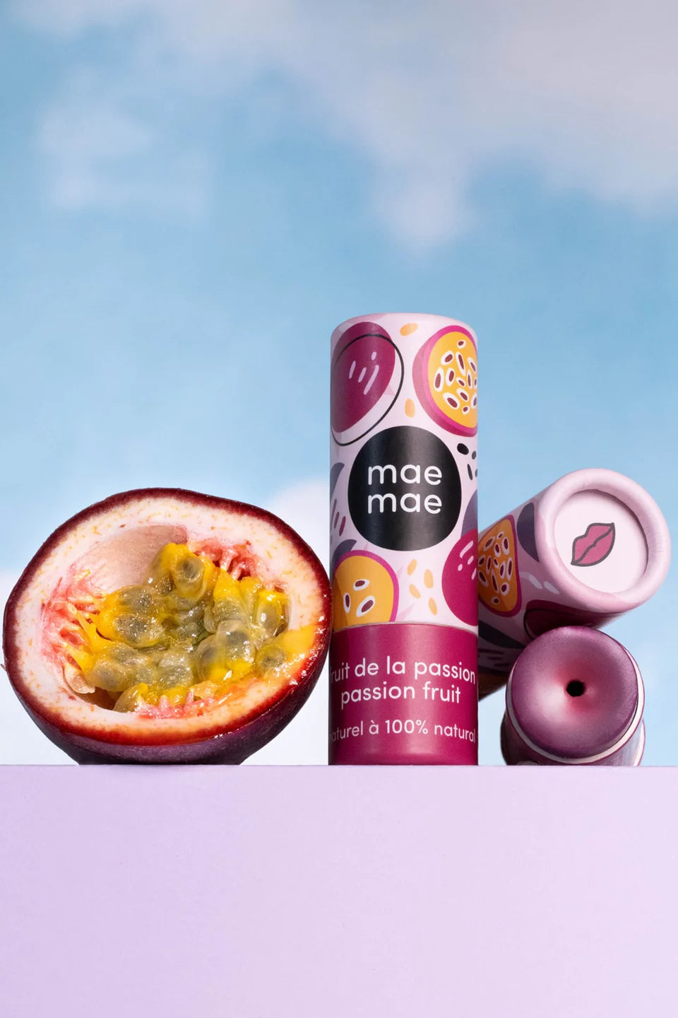 Baume à lèvres teinté naturel Mae Mae fruit de la passion présenté avec un fruit de la passion et emballages sur fond ciel bleu