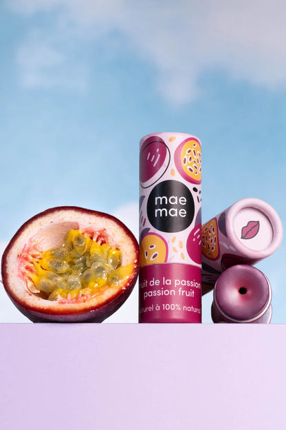 Baume à lèvres teinté naturel Mae Mae fruit de la passion présenté avec un fruit de la passion et emballages sur fond ciel bleu