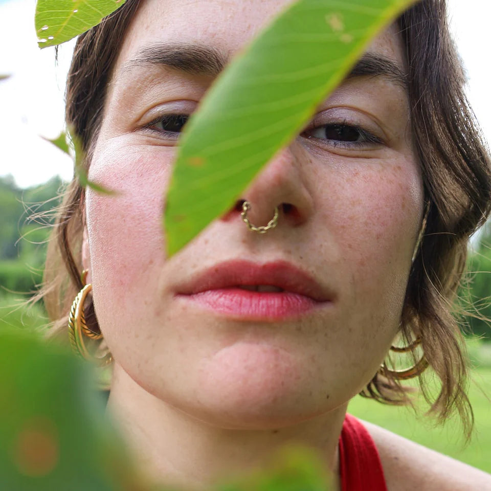 Close-up of a person’s face outdoors, partially framed by green leaves, wearing softly tinted red lips consistent with pomegranate lip balm.