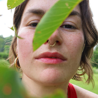 Close-up of a person’s face outdoors, partially framed by green leaves, wearing softly tinted red lips consistent with pomegranate lip balm.