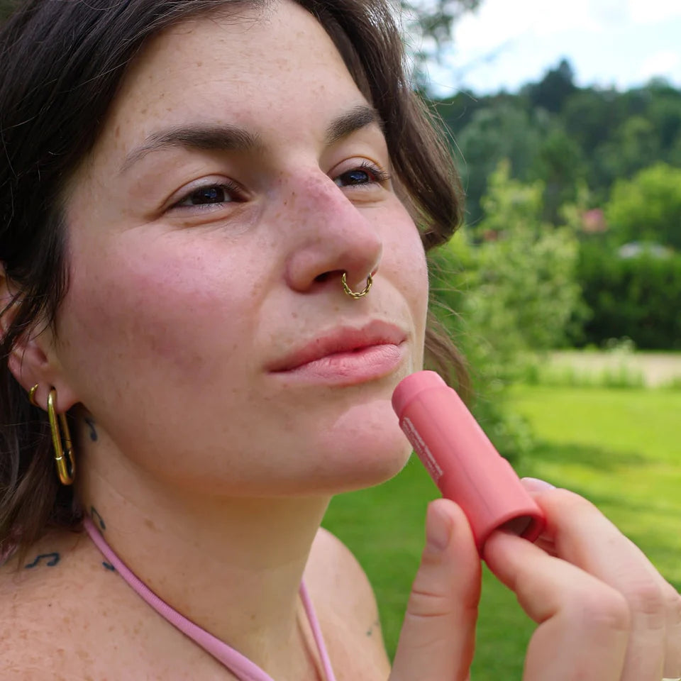 Application du baume à lèvres teinté à la pêche Mae Mae sur les lèvres en extérieur, offrant une teinte pêche naturelle dans un décor verdoyant.
